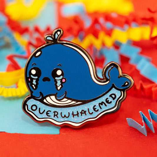 Close-up of the Overwhalemed Glittery Whale Enamel Pin by Innabox. The pin features a teary-eyed blue whale with a tiny water spout on its head, resting on a glittery blue wave with the word "Overwhalemed" written across it. Surrounded by colourful red, blue, and yellow crinkle paper.