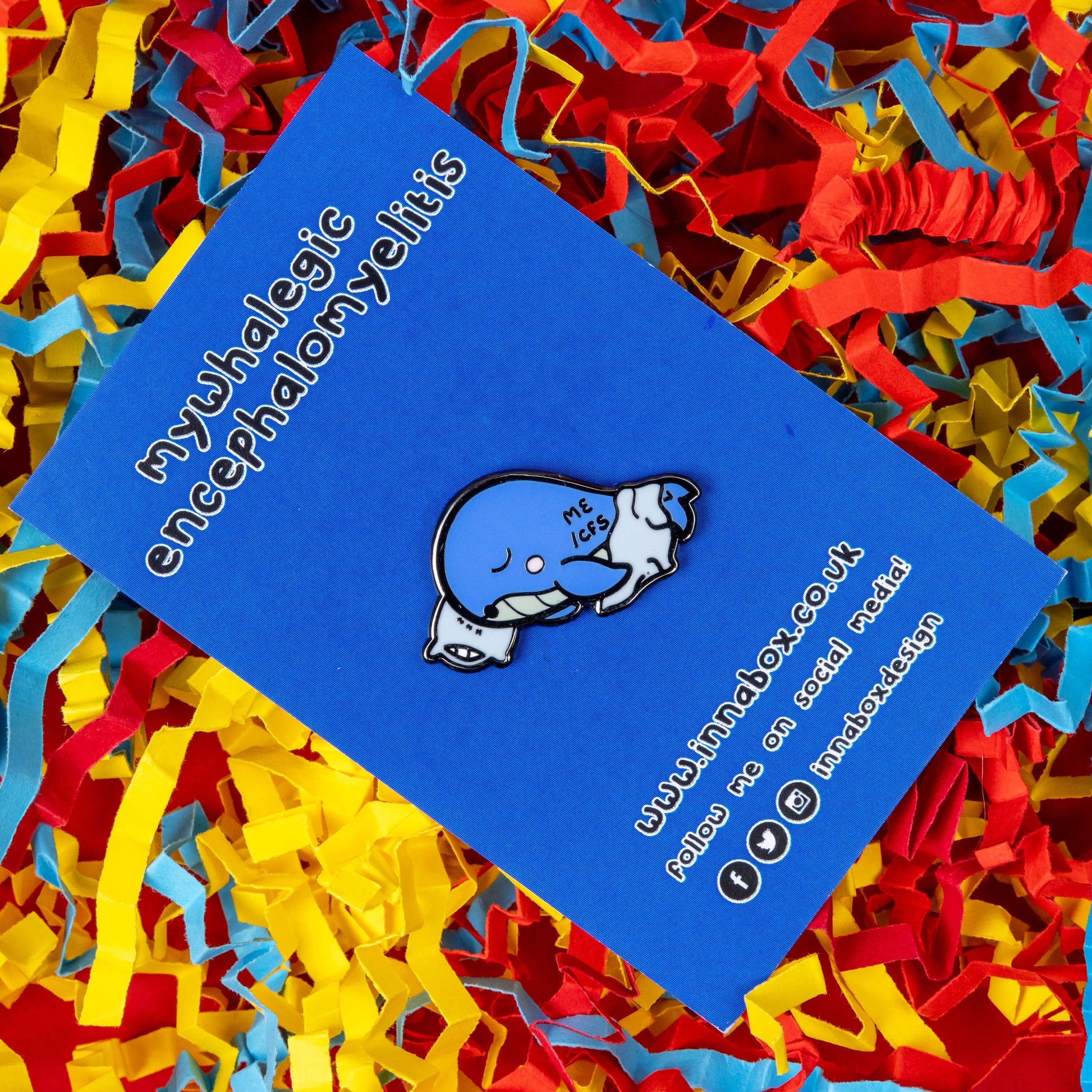 Mywhalegic Enamel Pin - Myalgic Encephalomyelitis (ME/CFS) on blue backing card on a red, blue and yellow coloured card confetti background. The enamel pin is a sleeping blue whale laying on a white pillow with a white blanket over it and ME/CFS written on its back. The enamel pin is designed to raise awareness for Myalgic encephalomyelitis or chronic fatigue syndrome