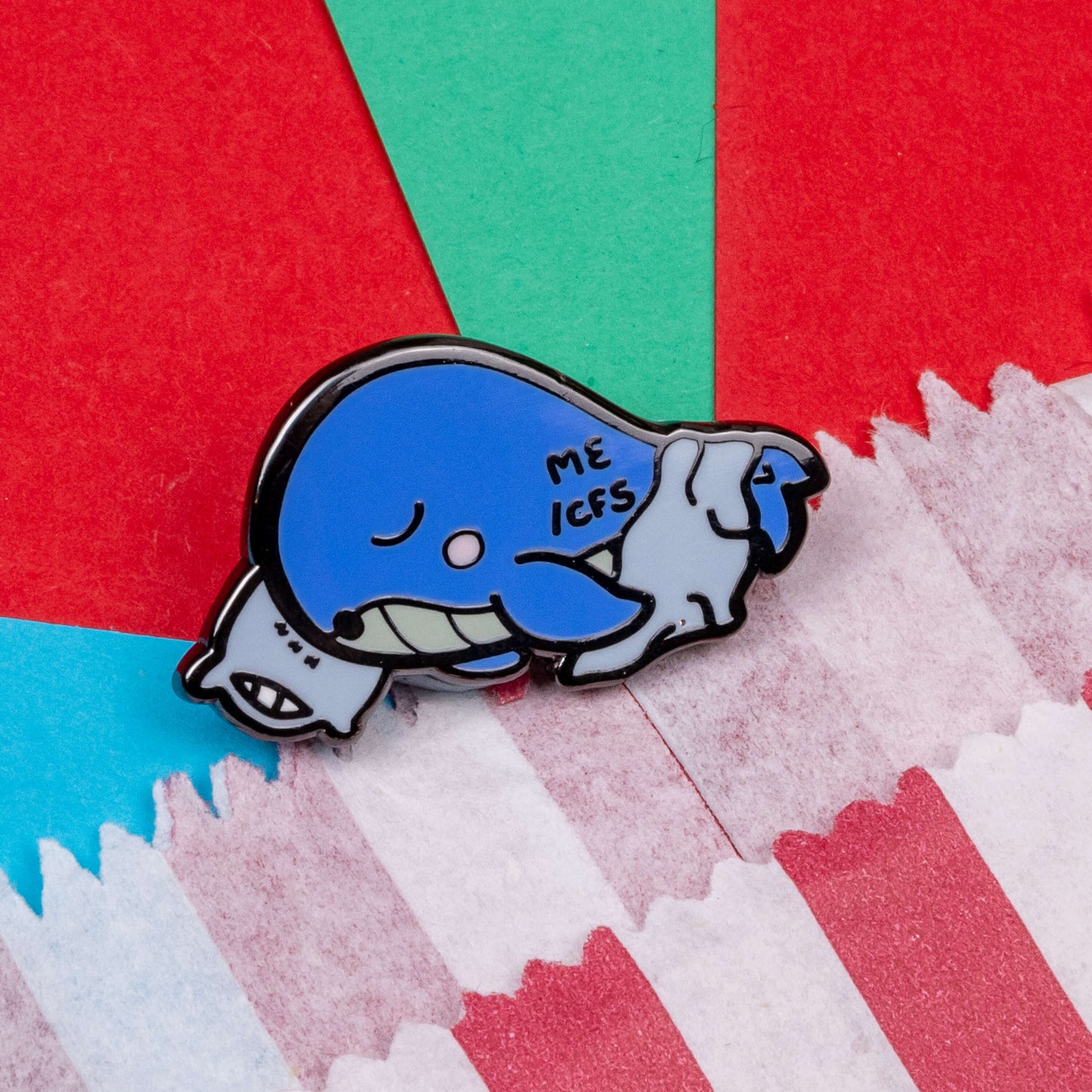 Mywhalegic Enamel Pin - Myalgic Encephalomyelitis (ME/CFS) on a red, blue and teal background next to a red and white striped paper bag. The enamel pin is a sleeping blue whale laying on a white pillow with a white blanket over it and ME/CFS written on its back. The enamel pin is designed to raise awareness for Myalgic encephalomyelitis or chronic fatigue syndrome