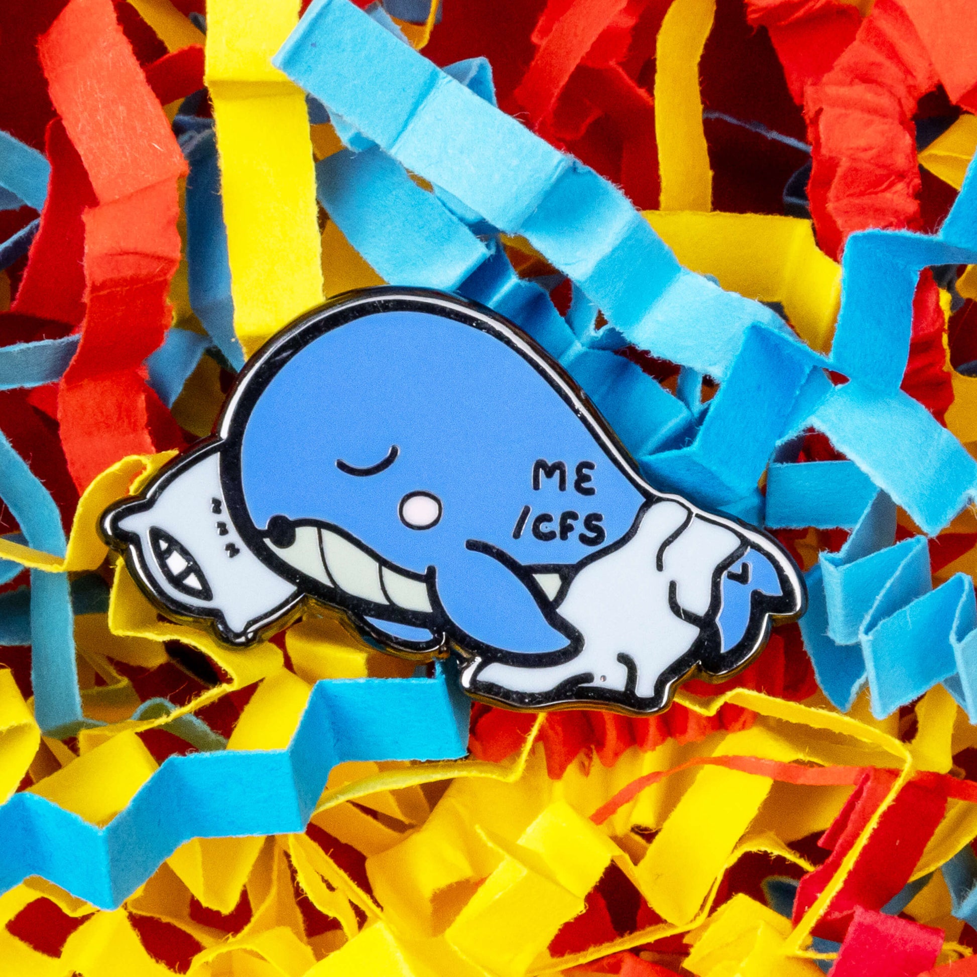 Mywhalegic Enamel Pin - Myalgic Encephalomyelitis (ME/CFS) on a blue, red and yellow card confetti background. The enamel pin is a sleeping blue whale laying on a white pillow with a white blanket over it and ME/CFS written on its back. The enamel pin is designed to raise awareness for Myalgic encephalomyelitis or chronic fatigue syndrome