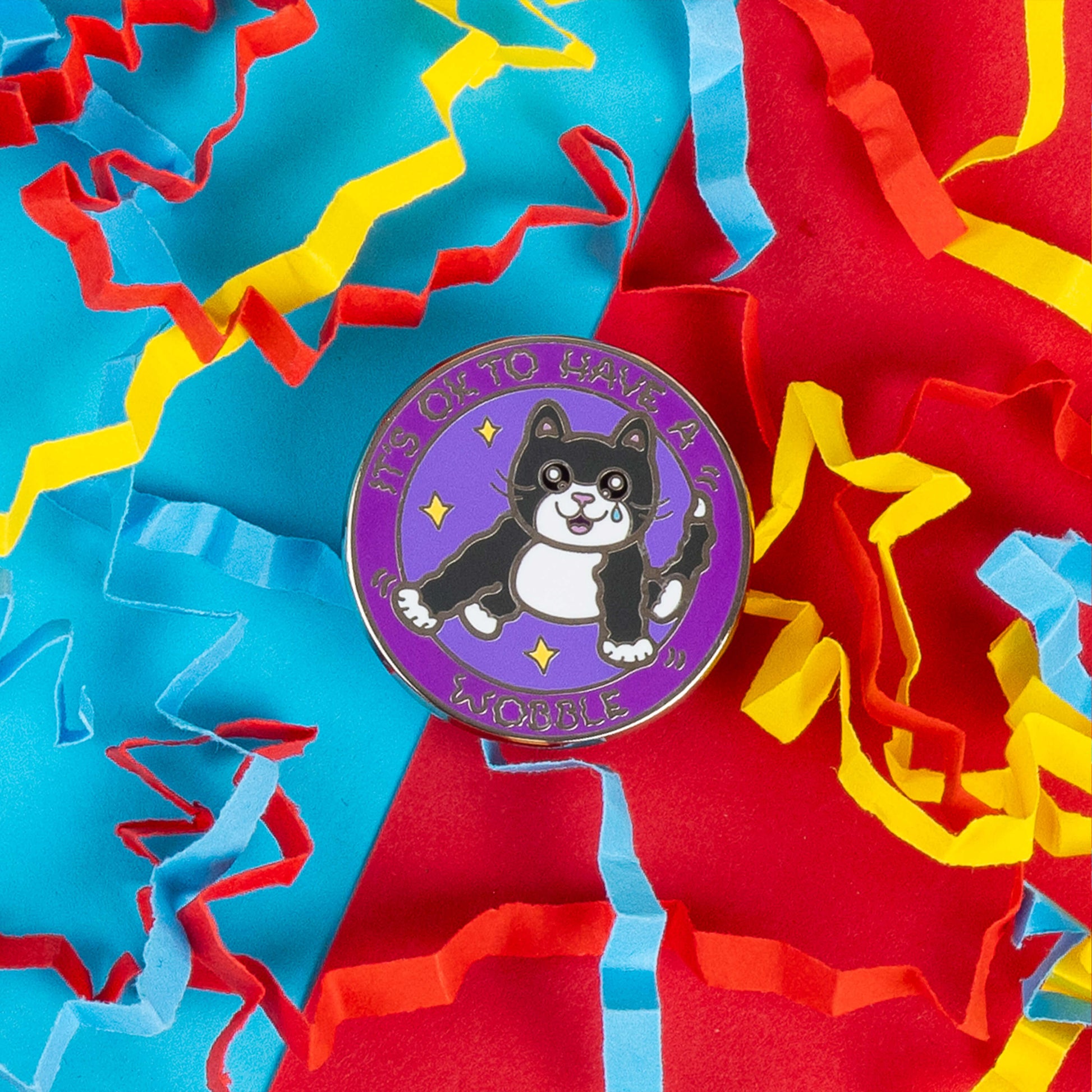 Round enamel pin by Innabox featuring an illustrated black and white cat with a tear in its eye, surrounded by stars. The text reads, "It's OK to Have a Wobble." The pin lies on a colourful backdrop of red, blue, and yellow paper confetti, giving it a vibrant and cheerful look.