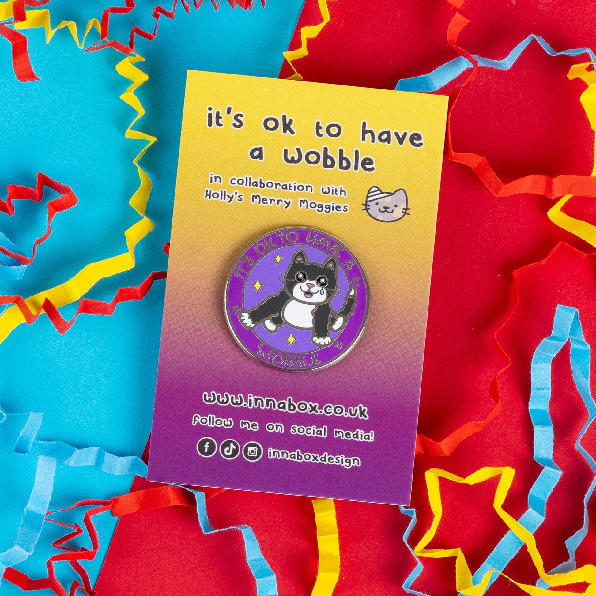 Enamel pin badge by Innabox with a design featuring a black and white cat with a tear, accompanied by the words, "It's OK to Have a Wobble." Displayed on a colourful backing card that says "In collaboration with Holly’s Merry Moggies," the pin is surrounded by colourful paper confetti.