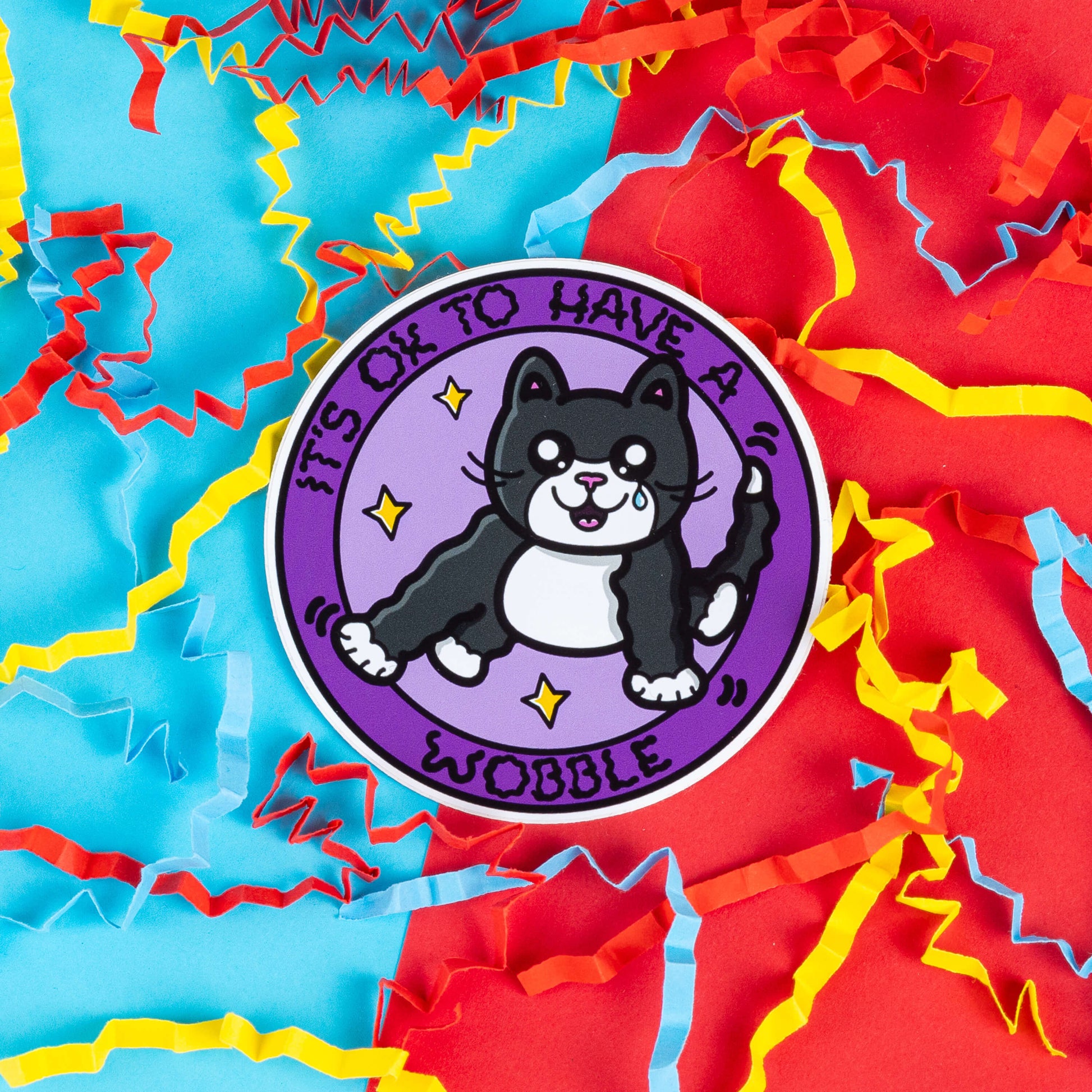 Round sticker by Innabox featuring an illustrated black and white cat with a tear in its eye, surrounded by stars. The text reads, "It's OK to Have a Wobble." The sticker lies on a colourful backdrop of red, blue, and yellow paper confetti, giving it a vibrant and cheerful look. Themed around mental and physical health with an underlying message to raise awareness for cerebellar hypoplasia in animals.