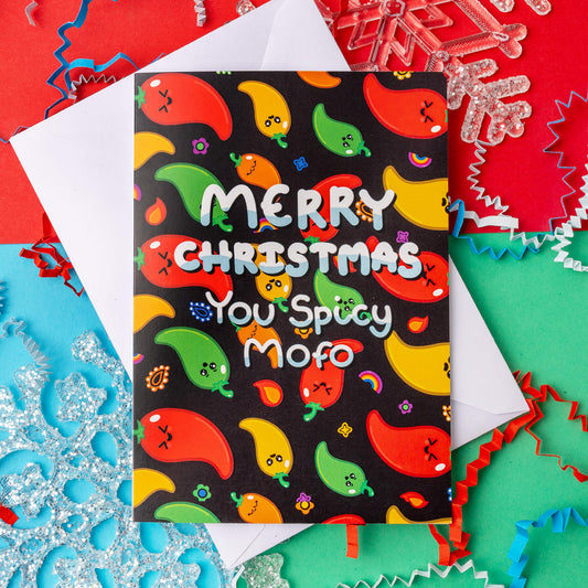 Merry Christmas you spicy mofo card by Innabox for spicy brain neurodivergent people. The card is covered in illustrated chillies in red, yellow, green, and orange with cute facial expressions, all set on a black background. Fun and bold lettering makes it a cheeky pun for those who are on the spectrum. Festive confetti and snowflakes surround the card.